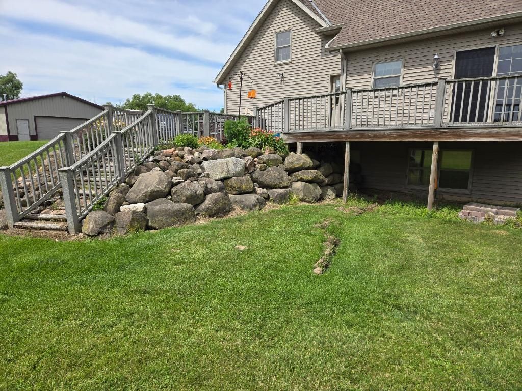 Before: aging boulder wall and worn grass slope beneath a raised wooden deck